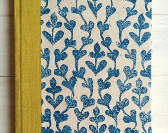 Op de afbeelding: Een rechthoekig notitieboek met een crèmekleurige kaft met een blauw bloemmotief. De rug en hoekaccenten zijn van mosterdgele stof. Het notitieboek is nieuw.