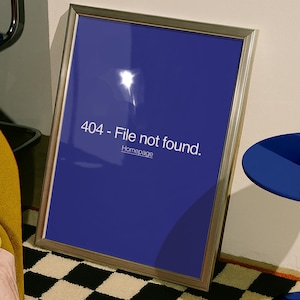 Puede incluir: Una impresión enmarcada con un marco plateado, que muestra un fondo azul oscuro con el texto "404 - File not found." y "Homepage." La impresión está apoyada contra una pared, junto a una mesa azul y una alfombra a cuadros.