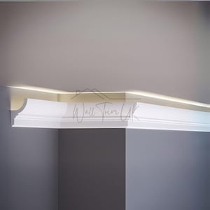 May include: White crown molding installed at the corner of a room. The molding has a curved design and is positioned against a light grey wall. The text "Wall Trim UK" is visible on the molding.