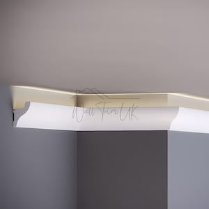 May include: A section of white crown molding installed at the corner of a room. The molding has a curved design and is positioned against a gray wall and a beige ceiling. The text "Wall Trim UK" is visible.