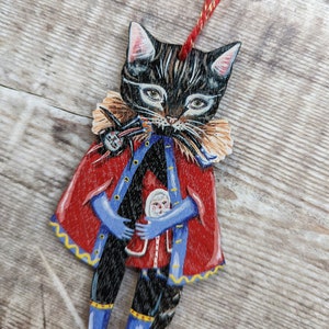 Peut inclure: Un chat noir portant un manteau rouge et bleu avec une bordure jaune tient une petite poup&eacute;e dans ses bras. Le chat a un visage blanc avec des yeux noirs et un nez noir. La poup&eacute;e porte une robe rouge et blanche et un chapeau bleu.