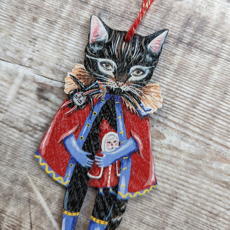 Peut inclure: Un chat noir portant un manteau rouge et bleu avec une bordure jaune tient une petite poup&eacute;e dans ses bras. Le chat a un visage blanc avec des yeux noirs et un nez noir. La poup&eacute;e porte une robe rouge et blanche et un chapeau bleu.