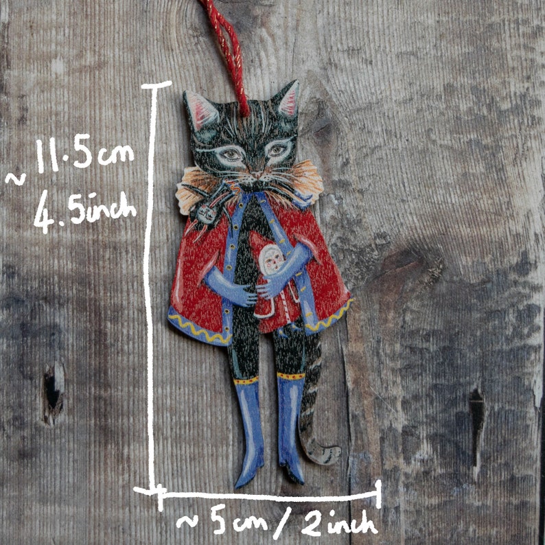 Peut inclure: Une d&eacute;coration de No&euml;l en bois en forme de chat avec une cape rouge et dor&eacute;e, des bottes bleues et une petite poup&eacute;e dans ses bras. La d&eacute;coration mesure environ 11,5 cm de haut et 5 cm de large.