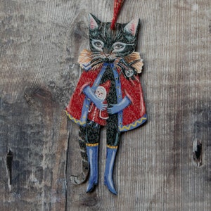 Peut inclure: Un chat noir portant un manteau rouge et bleu avec un col jaune et une petite poup&eacute;e dans ses bras. Le chat tient la poup&eacute;e avec sa patte droite. Le chat est un ornement de No&euml;l.