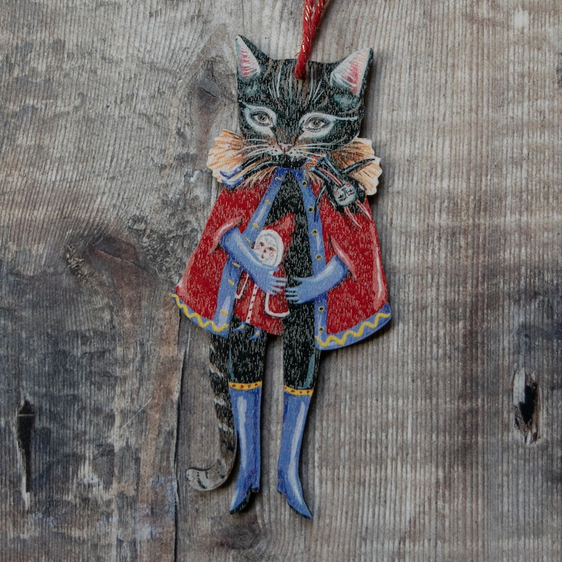 Peut inclure: Un chat noir portant un manteau rouge et bleu avec un col jaune et une petite poup&eacute;e dans ses bras. Le chat tient la poup&eacute;e avec sa patte droite. Le chat est un ornement de No&euml;l.