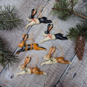May include: A selection of ceramic rabbit ornaments in black, orange, and brown, with gold string hangers. The rabbits are depicted in a running pose, with a red or green bow around their necks. The ornaments are arranged on a wooden surface with pine needles.
