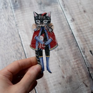 Peut inclure: Un chat noir habill&eacute; d'une cape rouge avec des bordures dor&eacute;es et des bottes bleues tient une petite poup&eacute;e dans ses bras. Le chat est une d&eacute;coupe en bois peinte avec des d&eacute;tails.