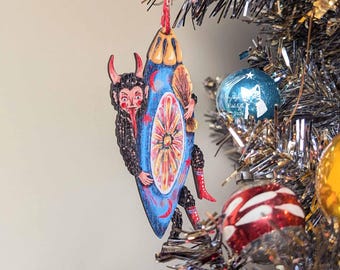 May include: A blue and red Christmas ornament featuring a cartoon devil holding a blue and red orb with a yellow flower design. The ornament is hanging on a silver tinsel Christmas tree.
