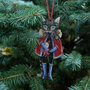 Peut inclure: Une d&eacute;coration de No&euml;l en bois en forme de chat noir portant une cape rouge et dor&eacute;e, un pantalon bleu et des bottes bleues. Le chat tient une petite poup&eacute;e dans ses bras.