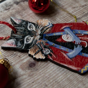 Peut inclure: Une d&eacute;coration de No&euml;l en bois en forme de chat noir portant un manteau rouge et bleu. Le chat tient une petite poup&eacute;e dans ses bras.