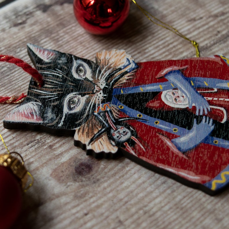 Peut inclure: Une d&eacute;coration de No&euml;l en bois en forme de chat noir portant un manteau rouge et bleu. Le chat tient une petite poup&eacute;e dans ses bras.
