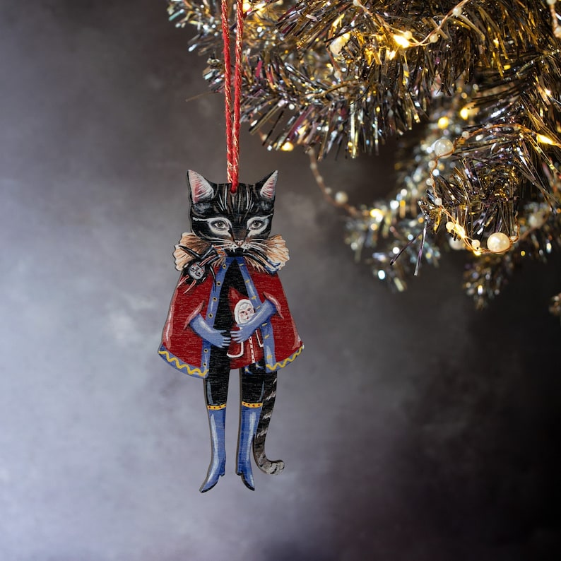 Peut inclure: Un chat noir portant une cape rouge et bleue et tenant une petite poup&eacute;e. Le chat est une d&eacute;coration de No&euml;l en bois peinte avec des d&eacute;tails.