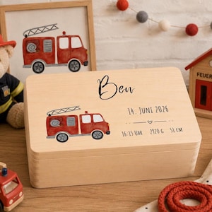 May include: A rectangular light-colored wooden keepsake box featuring a printed red fire truck illustration. The text reads Ben, 14. JUNI 2026, 16:15 UHR, 2920 G, and 51 CM. The box has a smooth finish and a hinged lid, displaying birth details.