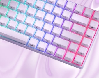 May include: White mechanical keyboard with a rainbow gradient key illumination. The word "keycap" is in purple text at the bottom of the image. The keyboard has a compact design. The keys are white.