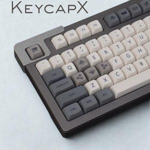May include: A mechanical keyboard with a gray and white color scheme. The keyboard features custom keycaps with unique symbols and the text "KEYCAPX" at the top. The keys are a mix of gray and off-white.