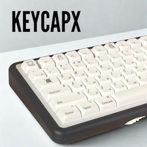 May include: A computer keyboard with white keycaps and black trim is shown. The keyboard has a standard QWERTY layout with symbols and letters. The word "KEYCAPX" is printed in large black letters at the top of the image.