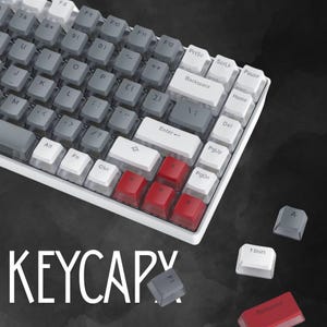May include: A white and gray mechanical keyboard with red accent keys. The keyboard has a variety of keys, including function keys, number keys, and letter keys. The word "KEYCAPY" is written in white letters at the bottom of the image.