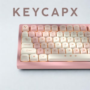 May include: A pink and white mechanical keyboard with the text "KEYCAPX" above it. The keyboard features a gradient colour scheme on the keycaps, transitioning from pink to white. The keys are arranged in a standard QWERTY layout.