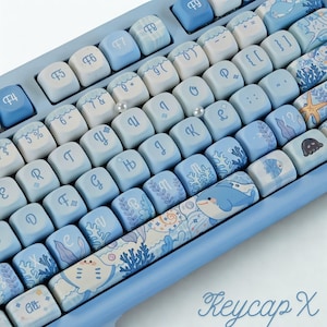 May include: A light blue mechanical keyboard with custom keycaps. The keycaps feature a variety of ocean-themed designs, including dolphins, coral, and other marine life. The keyboard has a light blue frame and the text "Keycap X" is visible.