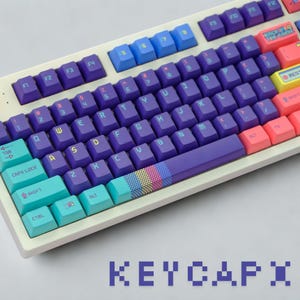 Peut inclure: Un clavier mécanique blanc avec des touches colorées. Les touches sont de différentes couleurs, notamment violet, bleu, rose et turquoise. Le clavier affiche le mot "KEYCAPX" en police pixelisée en bas.
