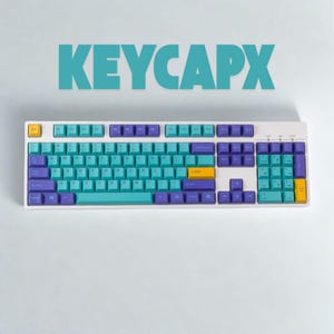 Puede incluir: Un teclado mecánico blanco con teclas turquesas, moradas y amarillas. El teclado tiene una distribución estándar con el texto "KEYCAPX" en turquesa en la parte superior. Las teclas están dispuestas en un patrón degradado.