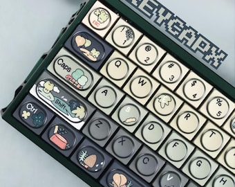 May include: A custom mechanical keyboard with green, gray, and cream-colored keycaps. The keycaps feature whimsical illustrations of plants and animals. The keyboard has the text "KEYCAPX" in the upper right corner.