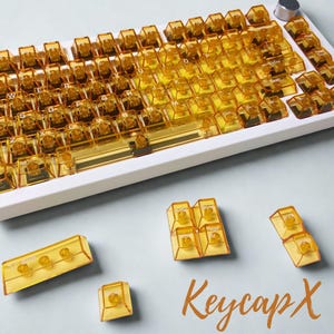 以下が含まれることがあります： 透明な琥珀色のキーキャップが付いた白いメカニカルキーボード。キーキャップは標準的なレイアウトで配置され、個々のキーキャップとキーキャップクラスターが個別に表示されます。「Keycap X」というテキストがオレンジ色の筆記体で書かれています。