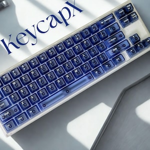 Può includere: Una tastiera con tasti blu e cornice bianca. I tasti sono traslucidi con lettere e simboli bianchi. La parola "Keycapx" è stampata in blu in alto a sinistra. La tastiera è su una superficie bianca.