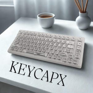 May include: A clear, transparent keyboard with white keys, resting on a white surface. The keyboard has a standard layout with letters, numbers, and function keys. The text "KEYCAPX" is printed in black below the keyboard.