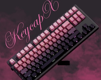 May include: A black mechanical keyboard with pink and purple keycaps. The keycaps transition from light pink to dark purple. The text "Keycap X" is written in pink cursive on the top left corner of the image.