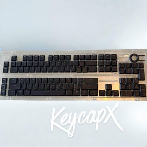 May include: A black computer keyboard featuring a full QWERTY layout, a numeric keypad, and five dedicated macro keys on the left side. The keyboard is encased in a clear, molded plastic protective cover and includes a circular keycap puller tool.