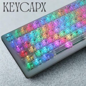 May include: A backlit keyboard with clear keycaps, illuminating in a rainbow of colors. The keyboard has a gray frame and the text "KEYCAPX" is visible in the upper left corner. The keys are labeled with letters, numbers, and symbols.