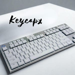 Puede incluir: Teclado de ordenador plateado y blanco con teclas blancas. El teclado tiene un diseño completo con teclas de función y un teclado numérico. La palabra "Keycapx" está escrita en cursiva negra encima del teclado.