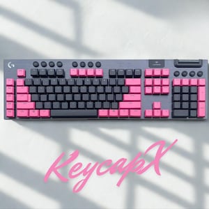 Puede incluir: Un teclado mecánico gris con teclas negras y rosas. El teclado tiene un teclado numérico y teclas de función. El texto "KeycapX" está escrito en rosa en cursiva debajo.