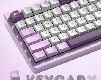 May include: A mechanical keyboard with a light purple base and keycaps in shades of white and purple. The keyboard features standard QWERTY layout with function keys and the brand name "KEYCAPX" in a stylized font.