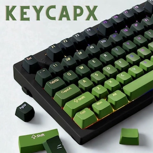May include: A black mechanical keyboard with green and black keycaps. The keyboard has the text "KEYCAPX" in green at the top. The keycaps are a mix of green and black, with white lettering. Several extra keycaps are scattered around the keyboard.