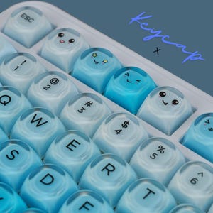 May include: Close-up of a keyboard with custom keycaps. The keycaps are a gradient of blue and white, with some featuring cute cartoon faces. The text "Keycap x" is written in a cursive font. The keys are arranged in a standard QWERTY layout.