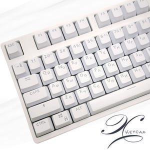 Puede incluir: Un teclado mecánico blanco con teclas blancas. El teclado presenta una distribución estándar con teclas alfanuméricas, teclas de función y teclas de control. Las teclas tienen caracteres en inglés y cirílico. El logotipo de la marca "KEYCAP" es visible en la esquina inferior derecha.
