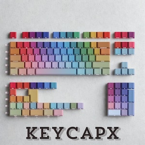 Puede incluir: Un teclado mecánico desmontado con teclas de colores arcoíris. Las teclas están dispuestas en filas y columnas, mostrando un degradado de colores que va del rojo al morado, azul y verde. El texto "KEYCAPX" está en la parte inferior.