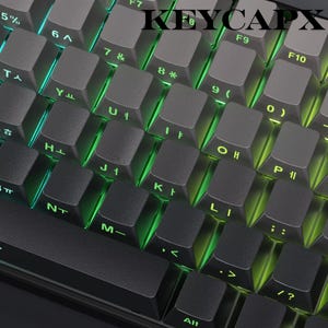 May include: Close-up of a black mechanical keyboard with gray keycaps. The keys are backlit with green LED lights. The word "KEYCAPX" is printed on the top of the keyboard.
