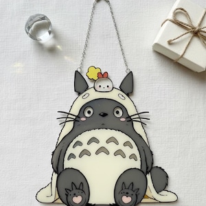 Może przedstawiać: Kapryśna, szaro-kremowa dekoracja wisząca inspirowana Totoro. Projekt przedstawia uśmiechniętą postać z małym pisklęciem na głowie, w komplecie z łańcuchem do zawieszenia. Dekoracja jest zaakcentowana czarnymi wąsami i wzorem w kształcie serca na stopach.