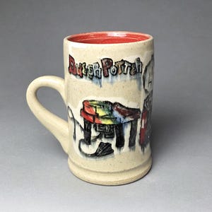 May include: A beige ceramic mug with a bright red interior. The exterior features a hand-painted illustration of a table and the text "Duesh Potten" in colorful, dripping lettering. The mug has a curved handle and a speckled, textured surface finish.