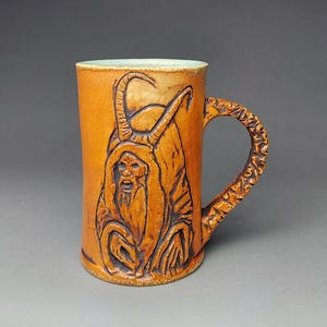 May include: An orange ceramic mug with a carved image of a hooded figure with horns. The handle has a textured, branch-like design. The interior rim has a light green glaze.