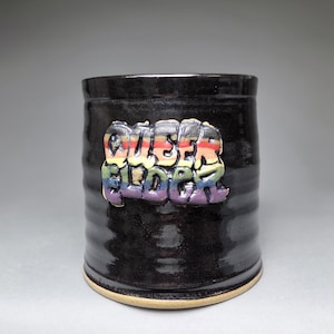 Puede incluir: Taza de cerámica negra con el texto "QUEER BLENDER" en colores arcoíris. La taza tiene forma cilíndrica con una superficie ligeramente texturizada y una base marrón claro. El texto está en relieve y parece pintado a mano.