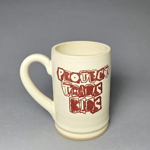 May include: Cream-colored ceramic mug with a curved handle. The mug features the red text "PROTECT TRANS KIDS" in a bold, blocky font. The mug has a slightly textured surface and a simple, cylindrical shape.