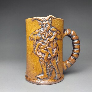 May include: A handmade ceramic mug featuring a detailed relief of a horned figure holding a child. The mug is a warm, earthy brown colour with a textured handle. A unique piece of art.