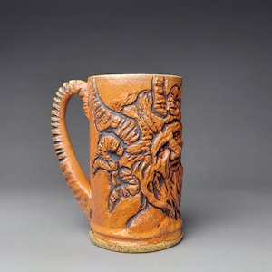 May include: An orange-brown ceramic mug with a detailed relief carving of a face and foliage. The handle has a textured, ridged design. The mug is suitable for hot or cold drinks.
