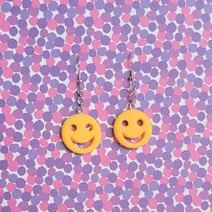 Peut inclure: Une paire de boucles d'oreilles smiley jaunes avec des crochets argentés. Les boucles d'oreilles sont rondes avec des yeux noirs et un sourire courbé. Le fond est un motif de points roses et violets.