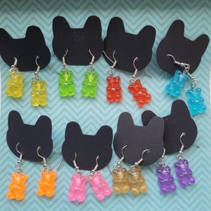 Könnte beinhalten: Eine Sammlung von Gummibärchen-Ohrringen in verschiedenen Farben, darunter Gelb, Grün, Rot, Blau, Orange, Rosa und Lila. Jedes Paar Ohrringe wird auf einer schwarzen Karte mit einem katzenförmigen Ausschnitt vor einem blau-weiß gemusterten Hintergrund präsentiert.