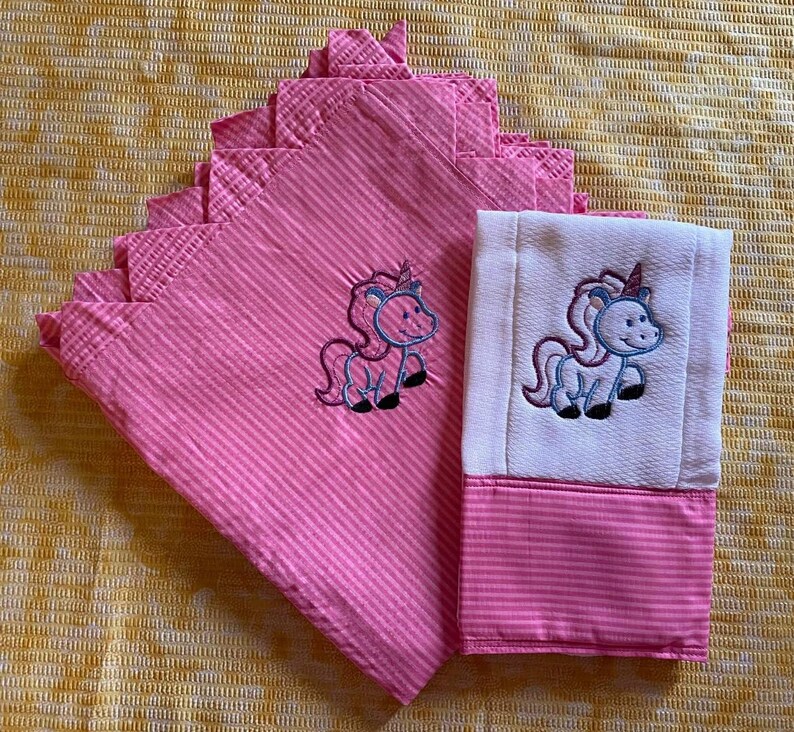 May include: Two pink and white striped fabric burp cloths with embroidered unicorn designs. The cloths are folded and stacked on top of each other.